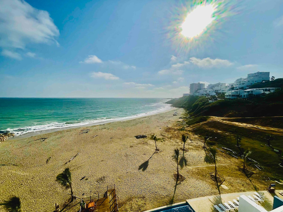 A panoramic view of a sandy beach meets the ocean, with gentle waves lapping at the shore. Sunlight creates a shimmering effect on the water, and the landscape is framed by palm trees. A few distant buildings line the hillside, adding depth to the scene.