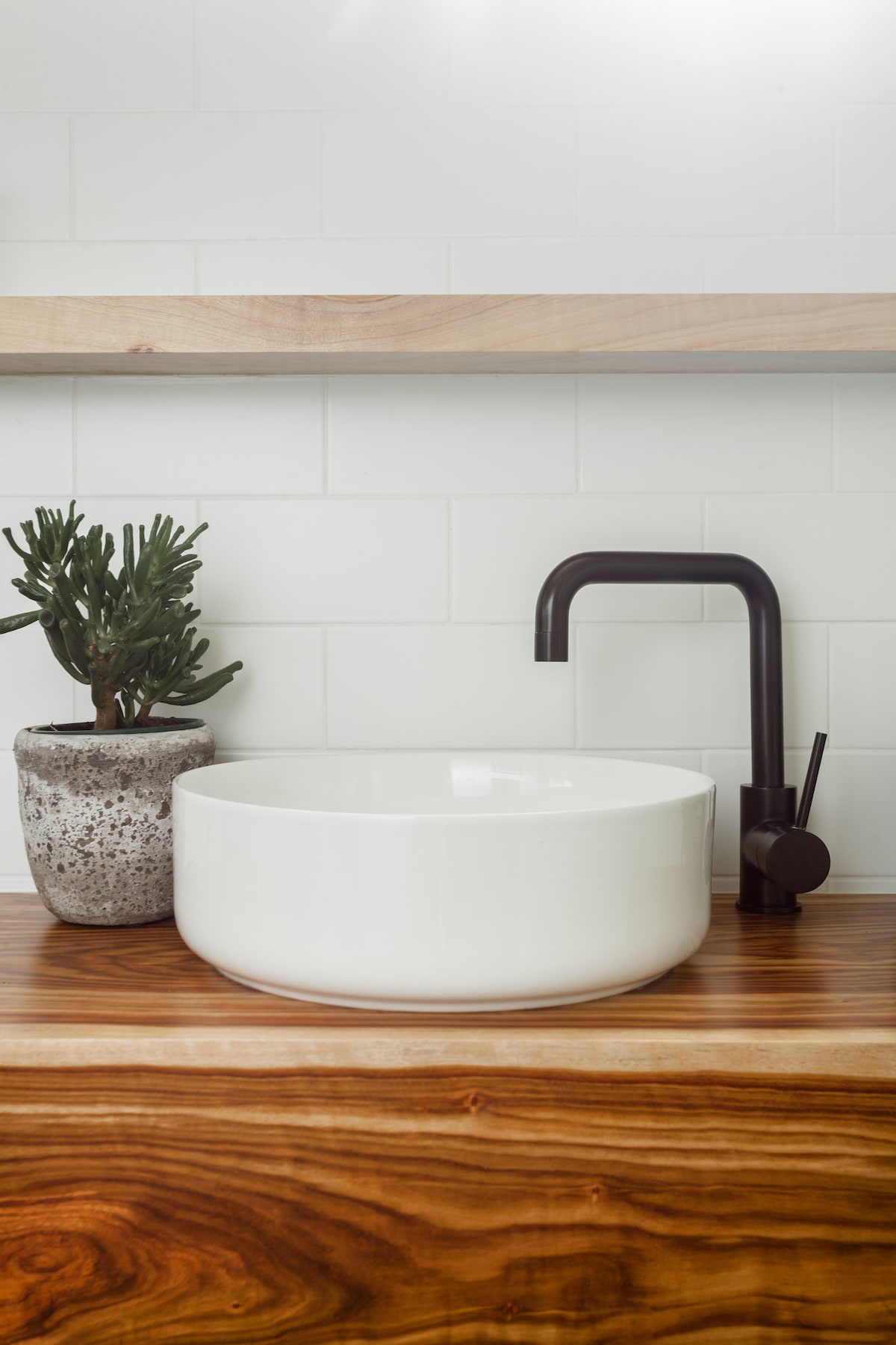 A modern bathroom sink is set on a wooden countertop featuring a rich grain. A circular white basin rests beside a small potted plant, while a sleek, dark faucet stands nearby, completing the minimalist aesthetic.