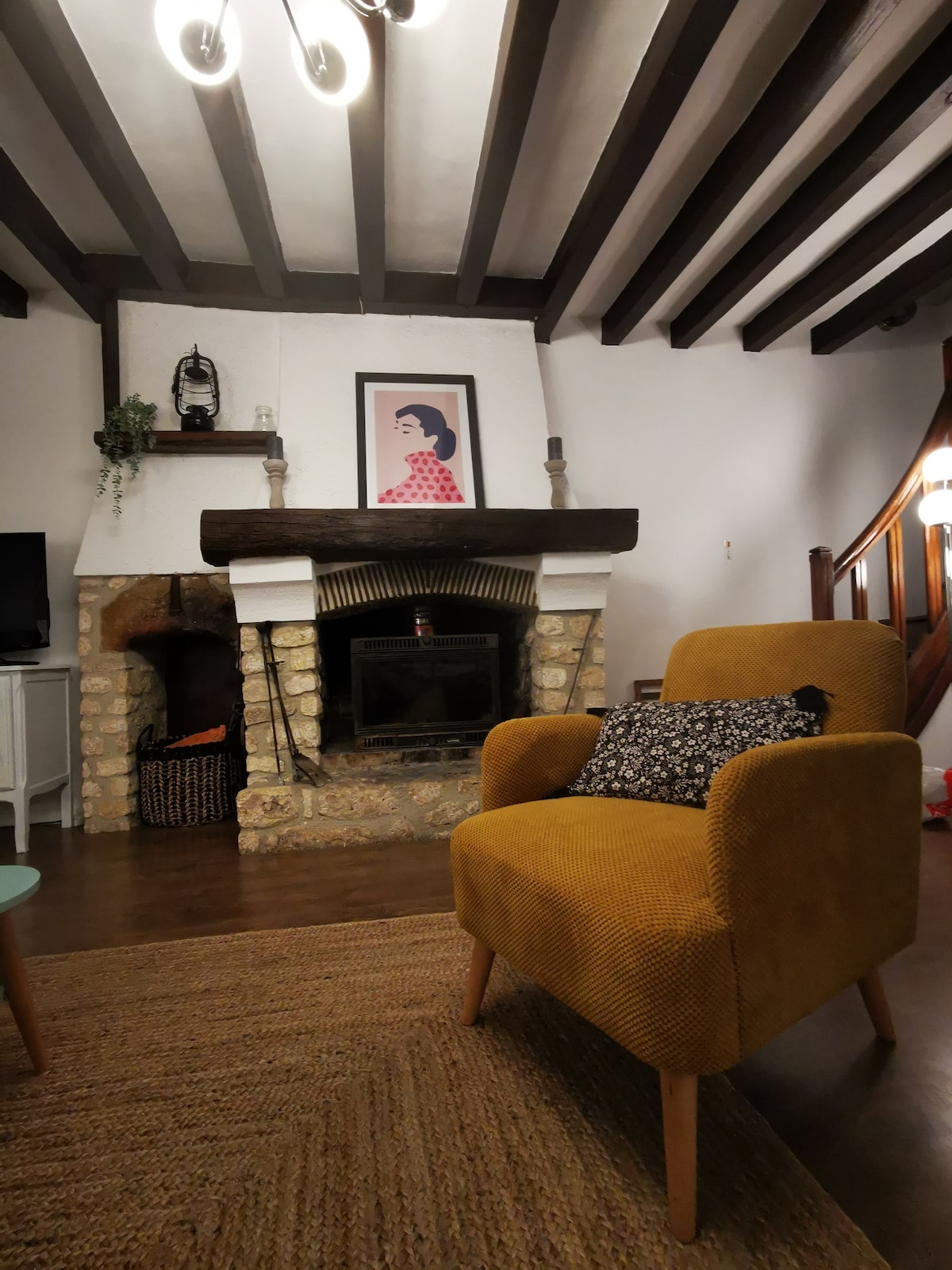 A cozy living area is highlighted by a soft yellow armchair with a patterned cushion, positioned near a rustic stone fireplace. Above the fireplace, a framed artwork adds a touch of character, while exposed wooden beams create a warm ambiance.