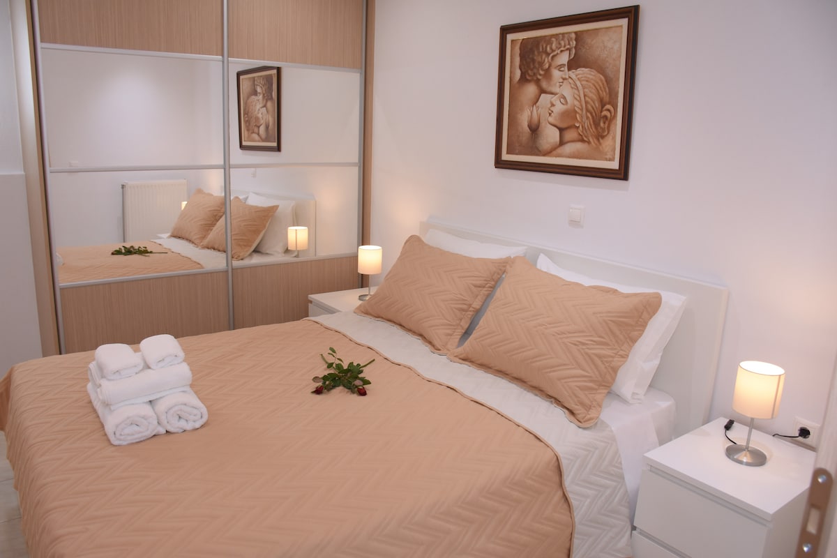 A well-appointed bedroom features a large bed dressed in neutral-toned linens, accented by matching throw pillows. A piece of artwork depicting a classical figure is displayed on the wall above the bed. Nightstands with lamps flank the bed, and a mirror reflects the serene layout.