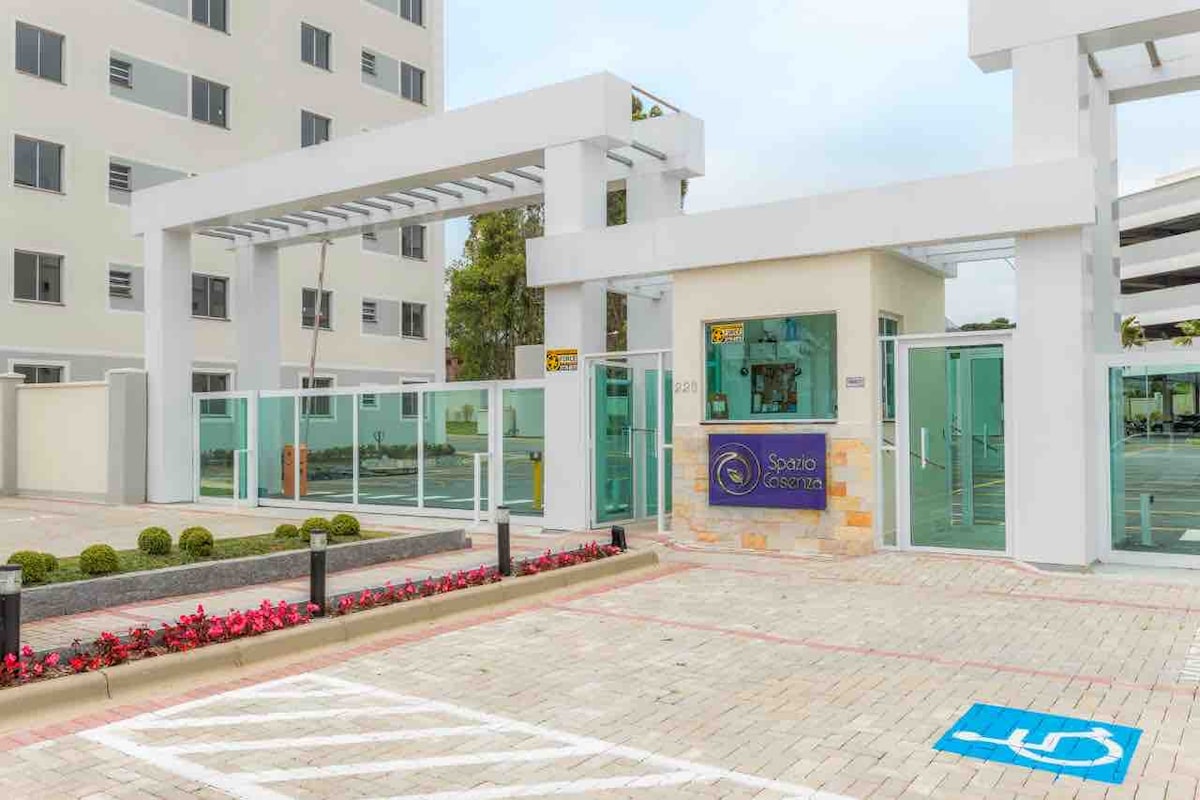 The entrance to the condominium is shown, featuring a modern structure with glass doors and a welcoming sign. Well-maintained landscaping includes low shrubs and vibrant flowers. Accessible parking spaces are visible, indicating thoughtful consideration for all guests. The overall design appears open and inviting.