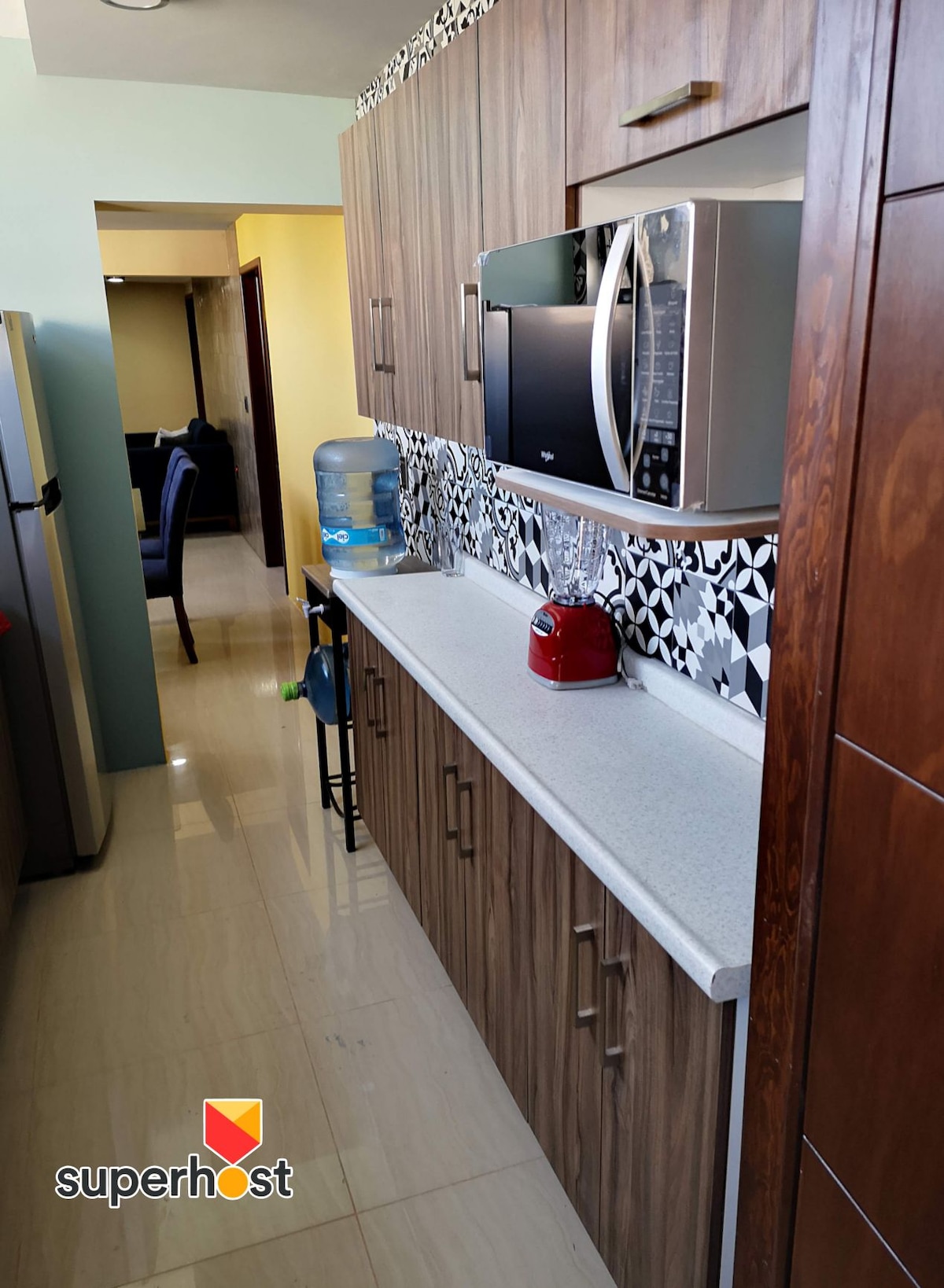 A modern kitchen is showcased, featuring wooden cabinetry and a light countertop. A microwave and a red kettle are displayed on the counter, with a water dispenser nearby. The wall behind the counter is decorated with black and white patterned tiles, enhancing the kitchen's design.