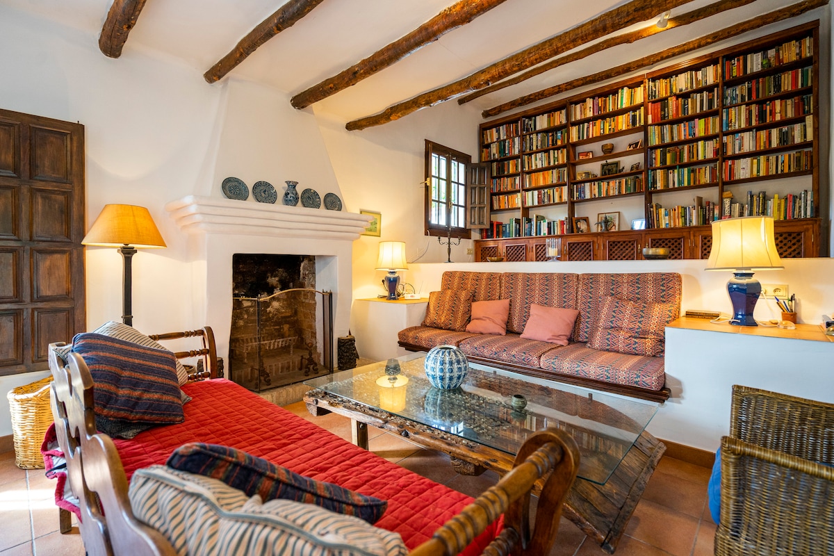 A cozy living room features a large bookshelf filled with books, a glass coffee table, and comfortable seating. A decorative lamp and two side tables provide soft lighting. The classic fireplace adds warmth, while natural light floods through the window with wooden shutters.