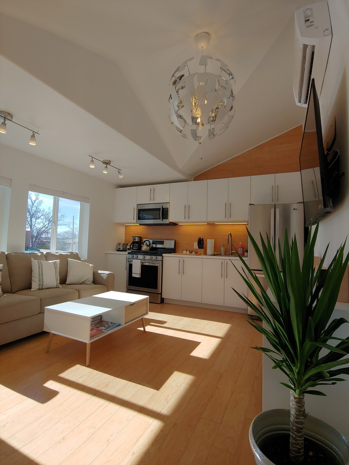 A spacious living area is featured, with a beige couch arranged around a white coffee table. Modern appliances are visible in the fully-equipped kitchen, along with a sleek dining area. Natural light floods the space through large windows, highlighting the contemporary design elements.
