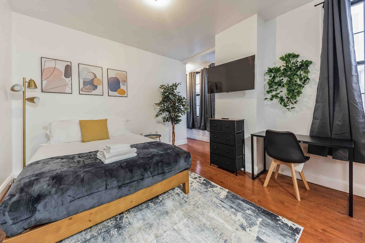 A spacious bedroom features a neatly made bed with a plush gray blanket and white linens. Modern art prints adorn the walls. A small desk is positioned beside a decorative plant, and a wall-mounted television is visible across from the bed.