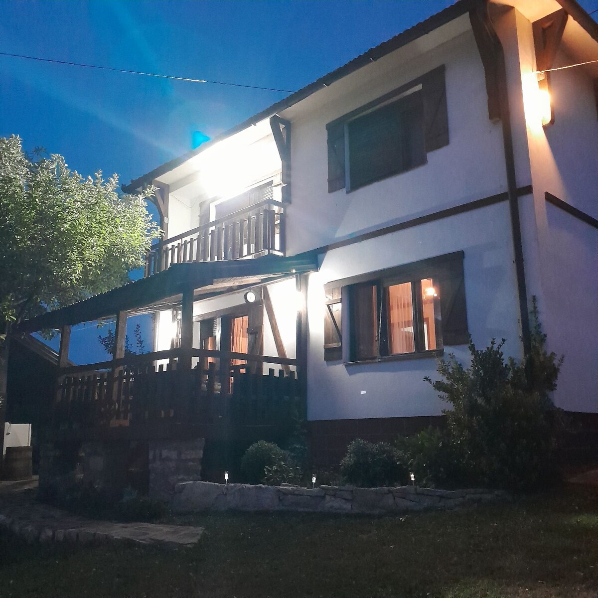 A two-story house is illuminated by exterior lights at night, highlighting its wooden balcony and decorative shutters. Windows are adorned with soft curtains, and landscaped greenery surrounds the property, providing a welcoming atmosphere.