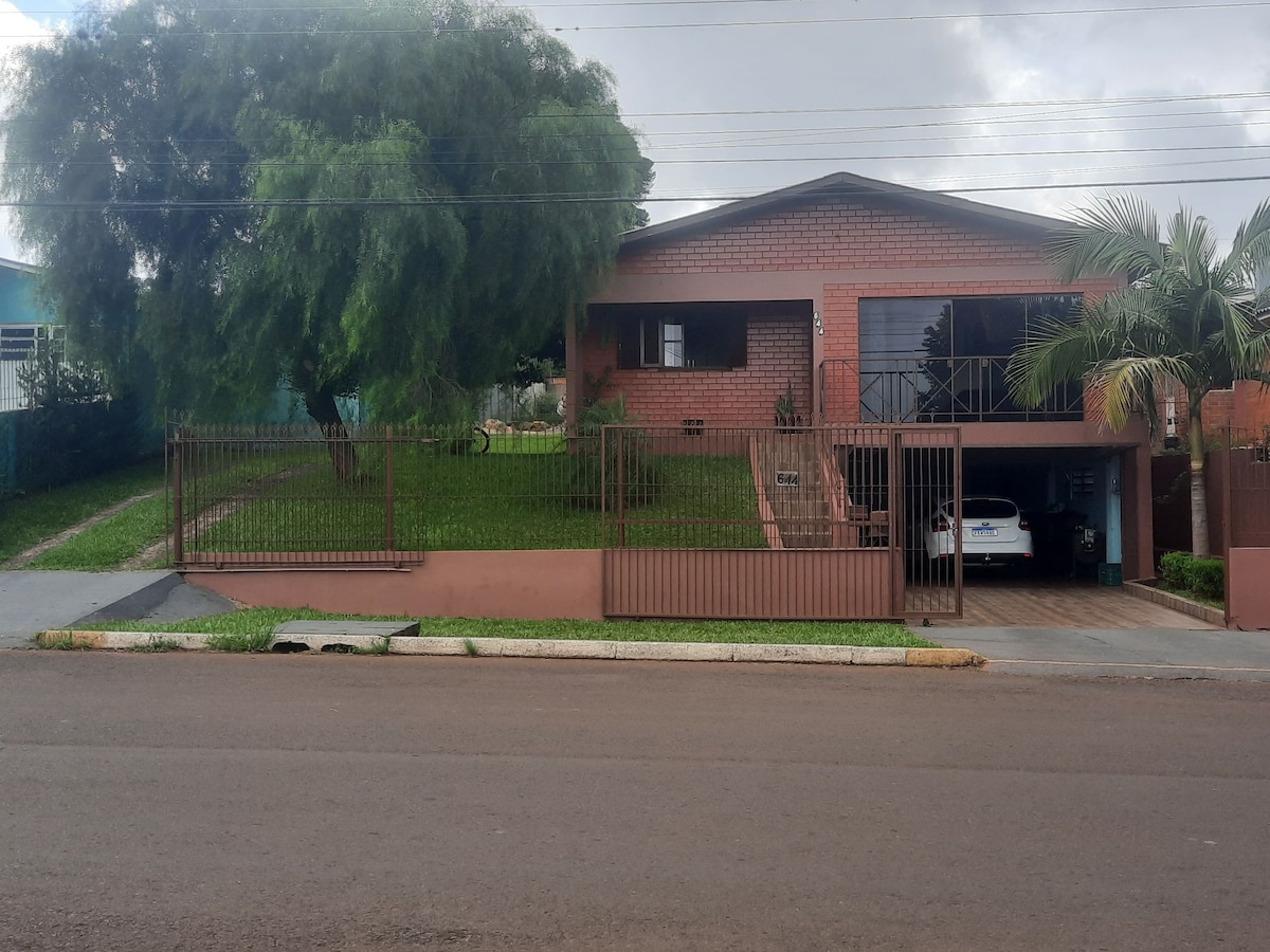 A two-story house is surrounded by a well-maintained lawn and trees. A gated driveway leads to a garage, with one vehicle visible inside. The brick exterior presents a warm tone, complemented by large windows that provide natural light. The home is set back from the street.