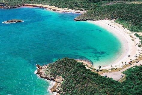 An aerial view of a serene beach with soft, sandy shores curving along crystal clear waters. The vibrant shades of blue blend seamlessly with the green hills in the background, creating a tranquil and inviting coastal landscape.