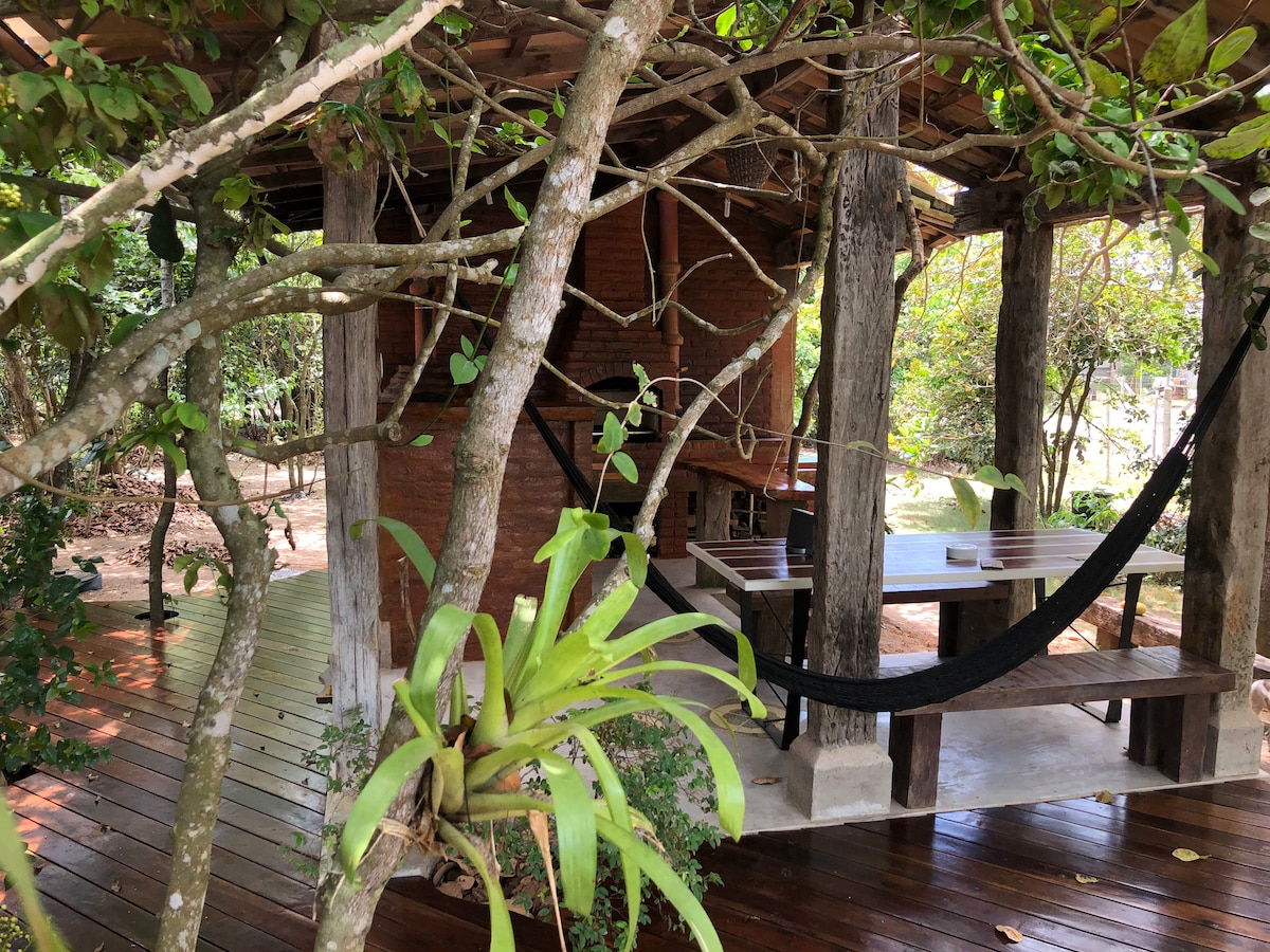 A rustic outdoor space features a wooden deck surrounded by greenery. A long table is positioned nearby, with a hammock suspended between sturdy posts. The structure behind has a brick façade and wooden beams, blending harmoniously with the natural surroundings.