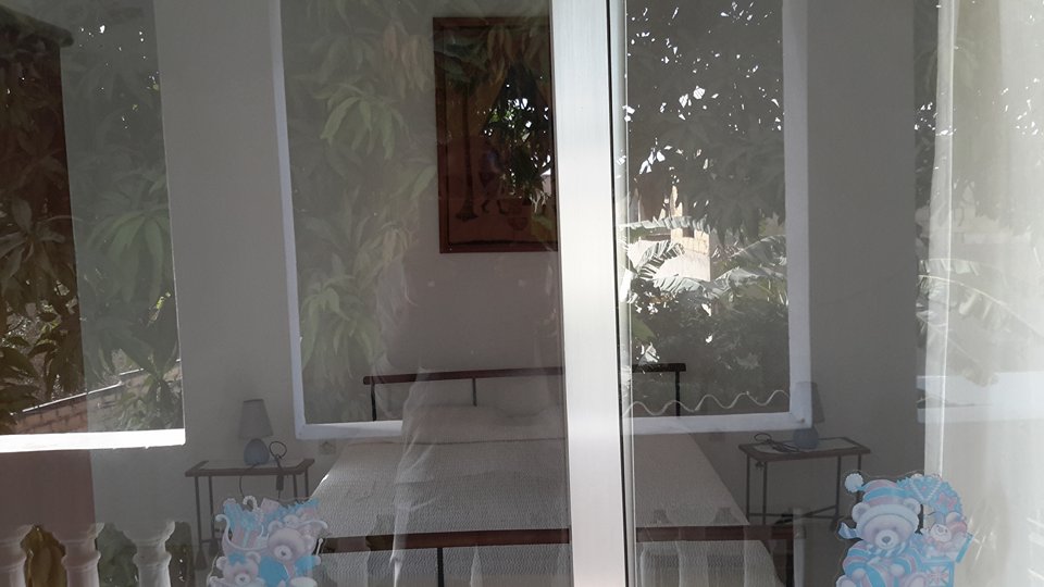 A peaceful bedroom is visible through a glass door, featuring a comfortable bed dressed in light-colored linens. Soft lighting fills the space, complemented by framed artwork on the wall. The view is framed by greenery, adding a touch of nature to the room.