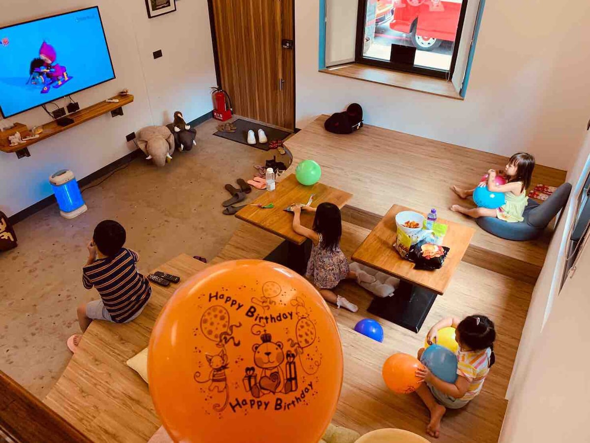 A spacious living area is shown, where children are engaged in activities while sitting on the floor. Colorful birthday balloons and snacks are scattered around, with a large screen displaying cheerful entertainment. Soft furnishings and natural light create a welcoming atmosphere.
