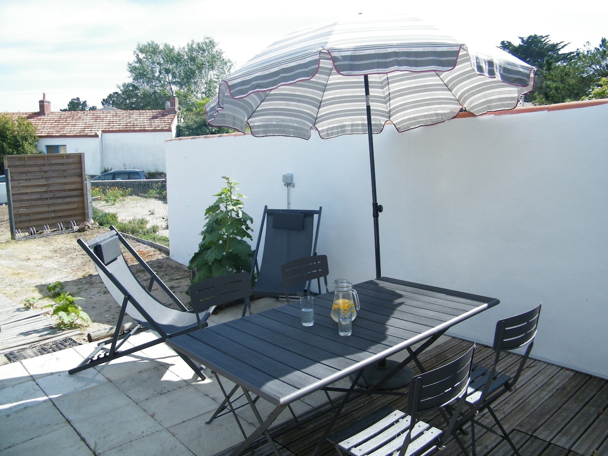 An outdoor terrace features a wooden table surrounded by four metal chairs. A large umbrella provides shade, while a lounge chair is positioned nearby. A small garden area is visible in the background, enhancing the outdoor setting.