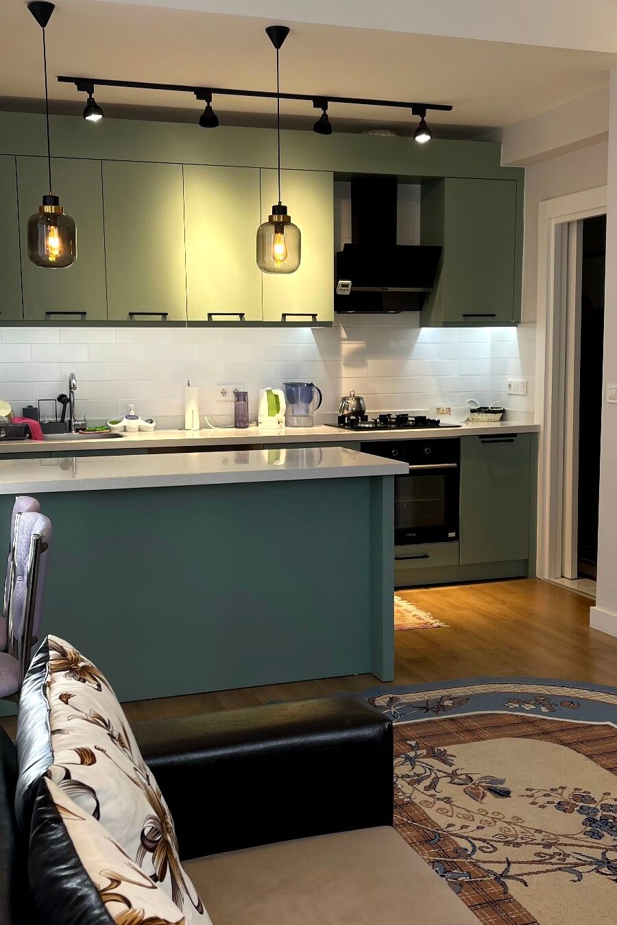A modern kitchen features green cabinets with a white tile backsplash. Pendant lights hang above the counter, illuminating the area. A stove and oven are integrated, alongside a countertop. A decorative rug lies on the floor, adding warmth to the space.