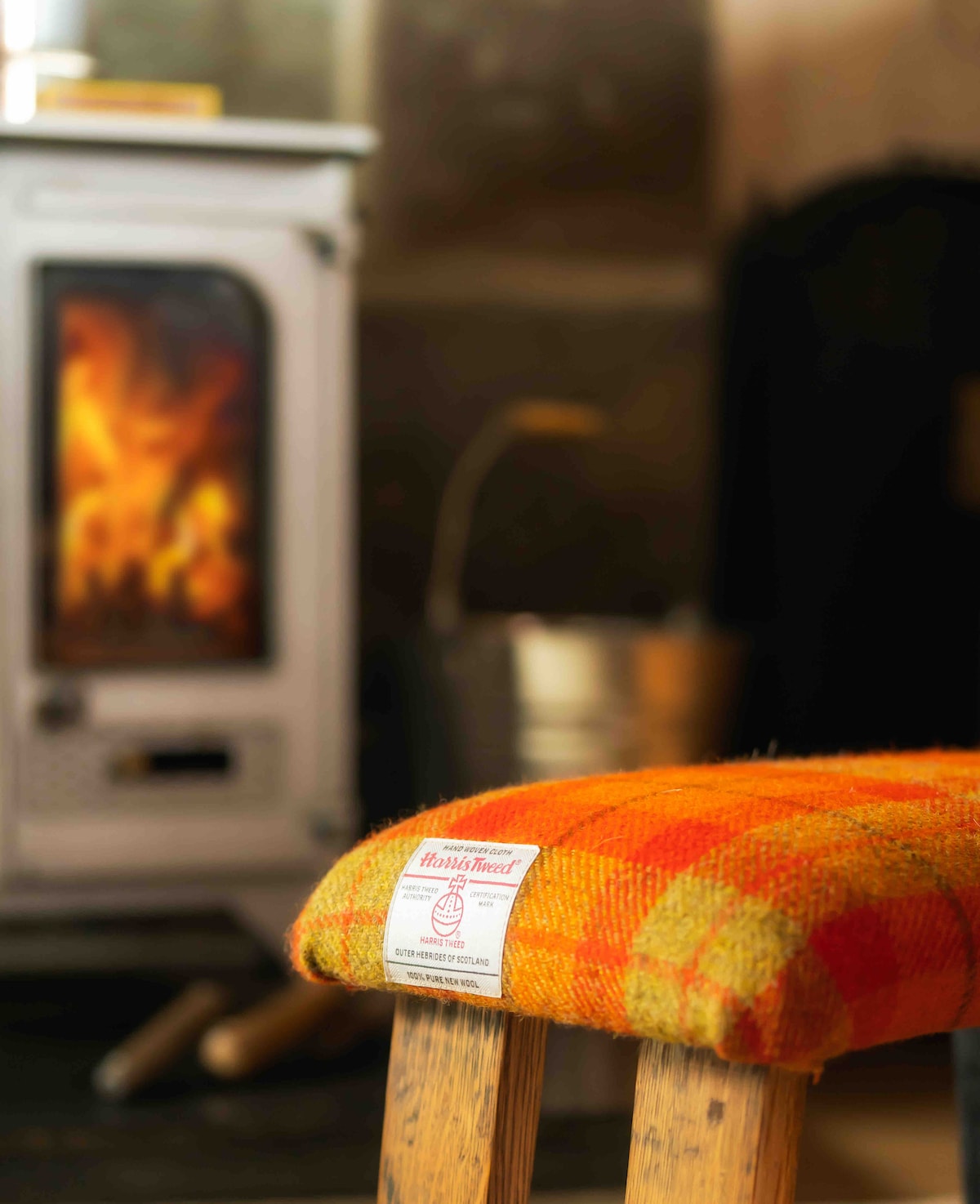 A wooden stool with a bright orange and yellow plaid cushioned seat is positioned near a traditional white wood-burning stove. The stove’s flame creates a warm glow in the background, while the surrounding area features a rustic decor atmosphere.