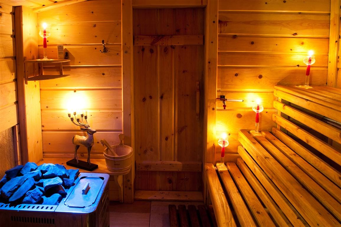 A wooden sauna interior is illuminated by soft light, highlighting the smooth wooden walls and benches. A stone heater is visible, accompanied by a wooden bucket and ladles. The door remains closed, providing a sense of privacy and warmth within the rustic space.