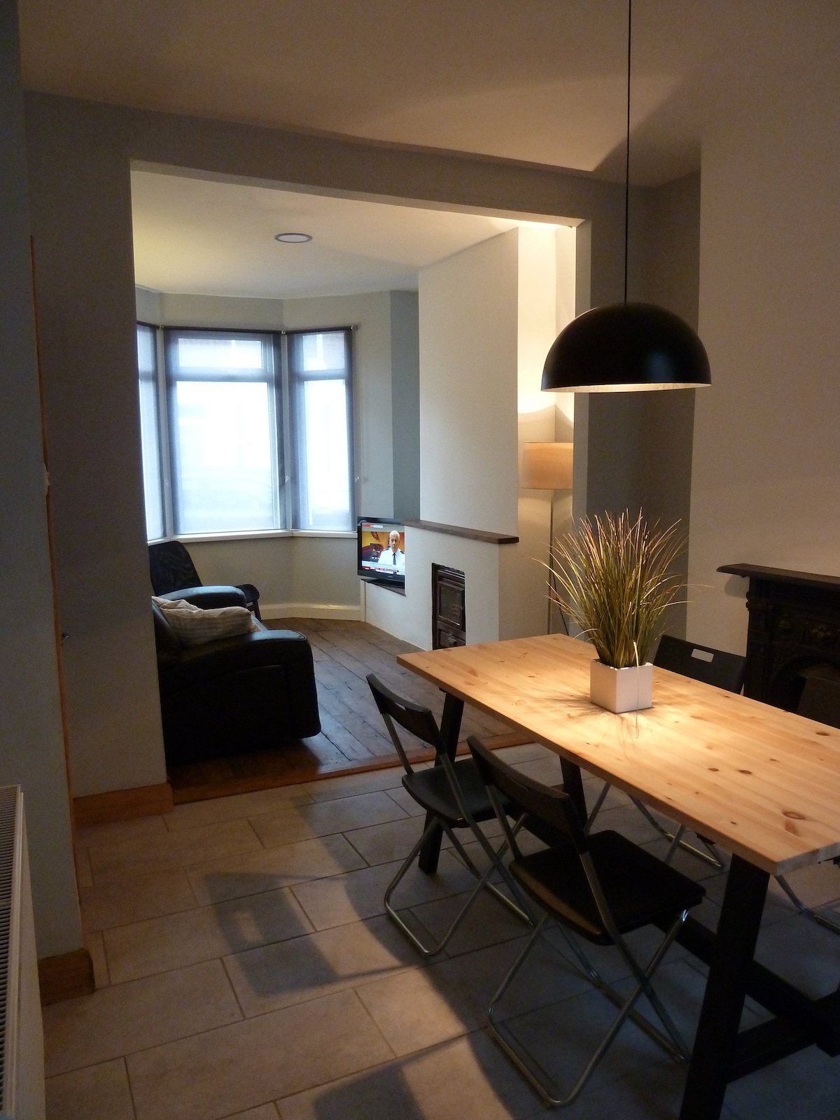 A dining area is shown with a wooden table and four folding chairs, illuminated by a pendant light. A cozy lounge nook is visible in the background, featuring a black armchair and a television mounted on the wall. Natural light streams through the bay window.