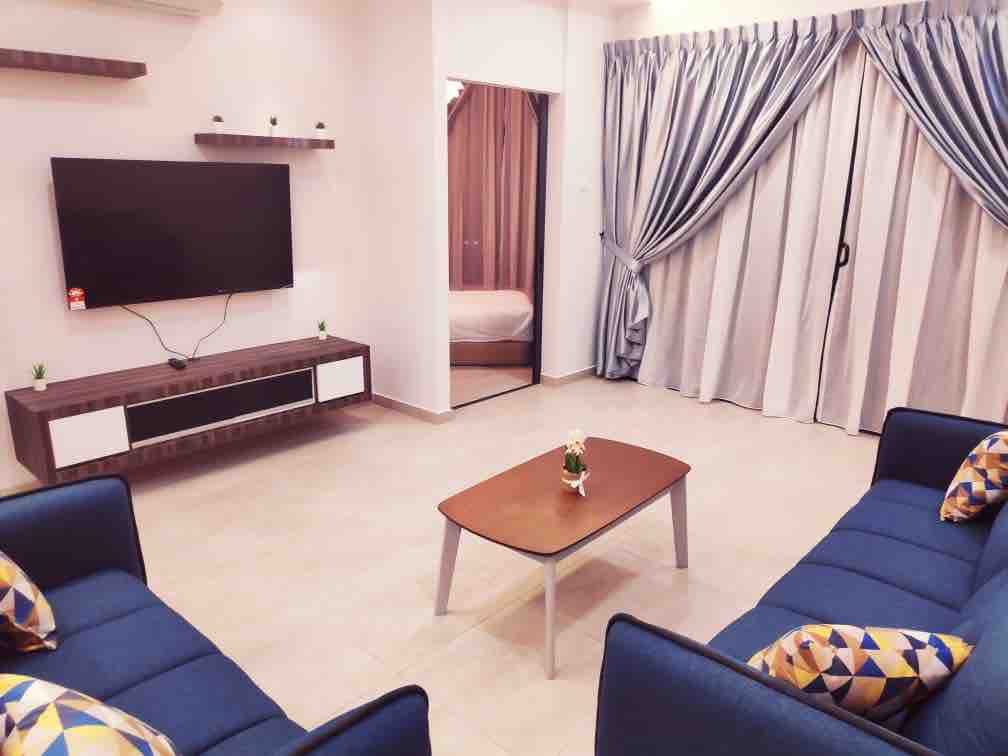 A spacious living room is revealed, featuring a large wall-mounted TV above a modern entertainment unit. Comfortable blue sofas are arranged around a stylish coffee table, adorned with decorative plants. Light-colored curtains frame the windows, allowing soft natural light to enter the space.