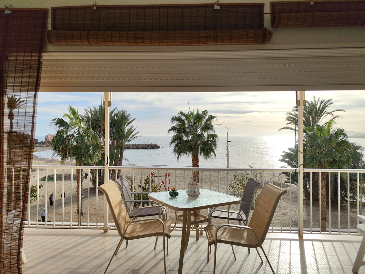 A spacious terrace is shown with two light chairs and a round table, offering views of palm trees and the beachfront. Sunlight reflects off the water, creating a serene atmosphere. Shades partially cover the windows, enhancing the overall tranquility of the space.