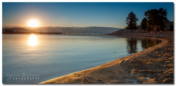 A serene lakeshore reflects the golden hues of a sunrise over calm waters. The sandy beach curves gently along the lake's edge, while silhouettes of distant trees and a dock can be seen in the background, enhancing the tranquility of the scene.