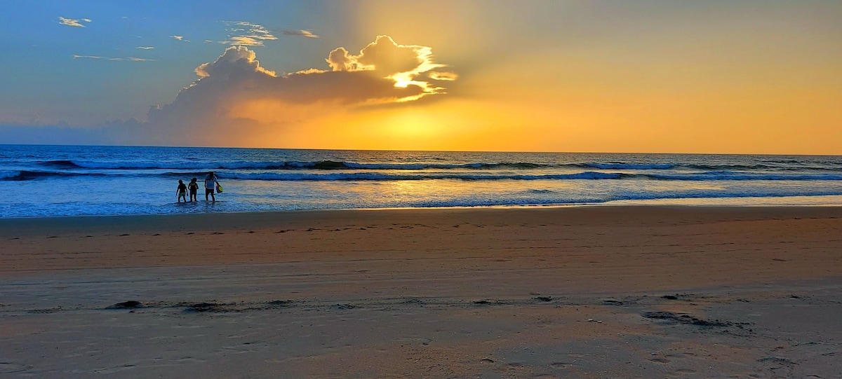 A serene beach scene is depicted with gentle waves lapping at the shore. Silhouettes of individuals are visible wading into the water against a backdrop of a vibrant sunset, casting soft light across the ocean surface.