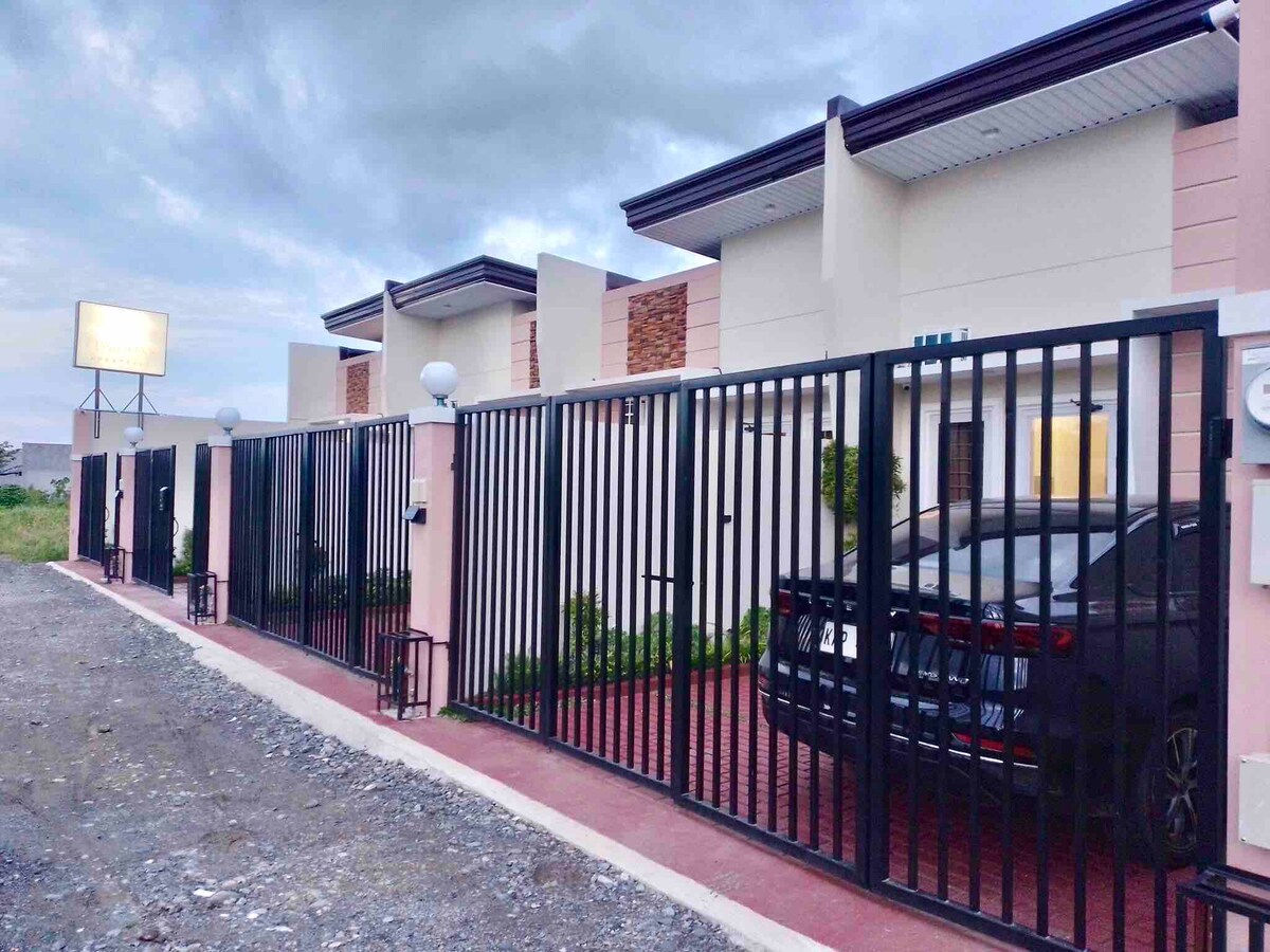 A gated entrance features a well-maintained exterior with a paved pathway leading to the apartments. A small car is parked within the private parking area, and outdoor lighting is positioned along the fence. The building exhibits modern architectural elements with a neutral color palette.