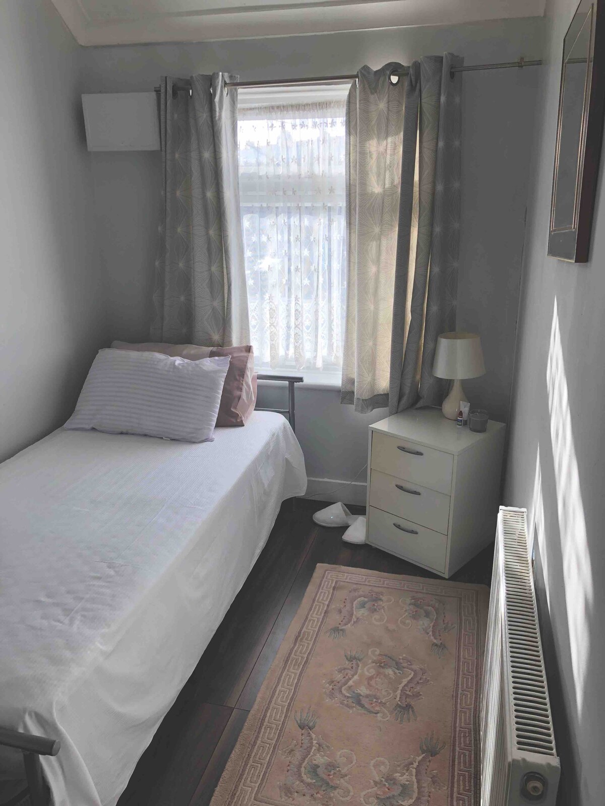 A cozy bedroom is defined by a single bed dressed in white linens and accented with a couple of pillows. Natural light filters through textured curtains, casting soft shadows. A small nightstand is beside the bed, and a lamp provides additional illumination, with a rug adding warmth to the wooden floor.