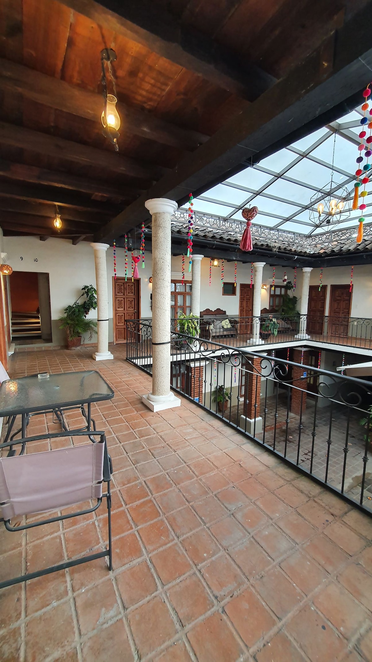 A balcony area with a tiled floor is framed by wooden beams and decorative pendant lights. An open space overlooks a central courtyard adorned with plants. Multiple doors lead into nearby rooms, while colorful decorations lend a vibrant touch to the space.