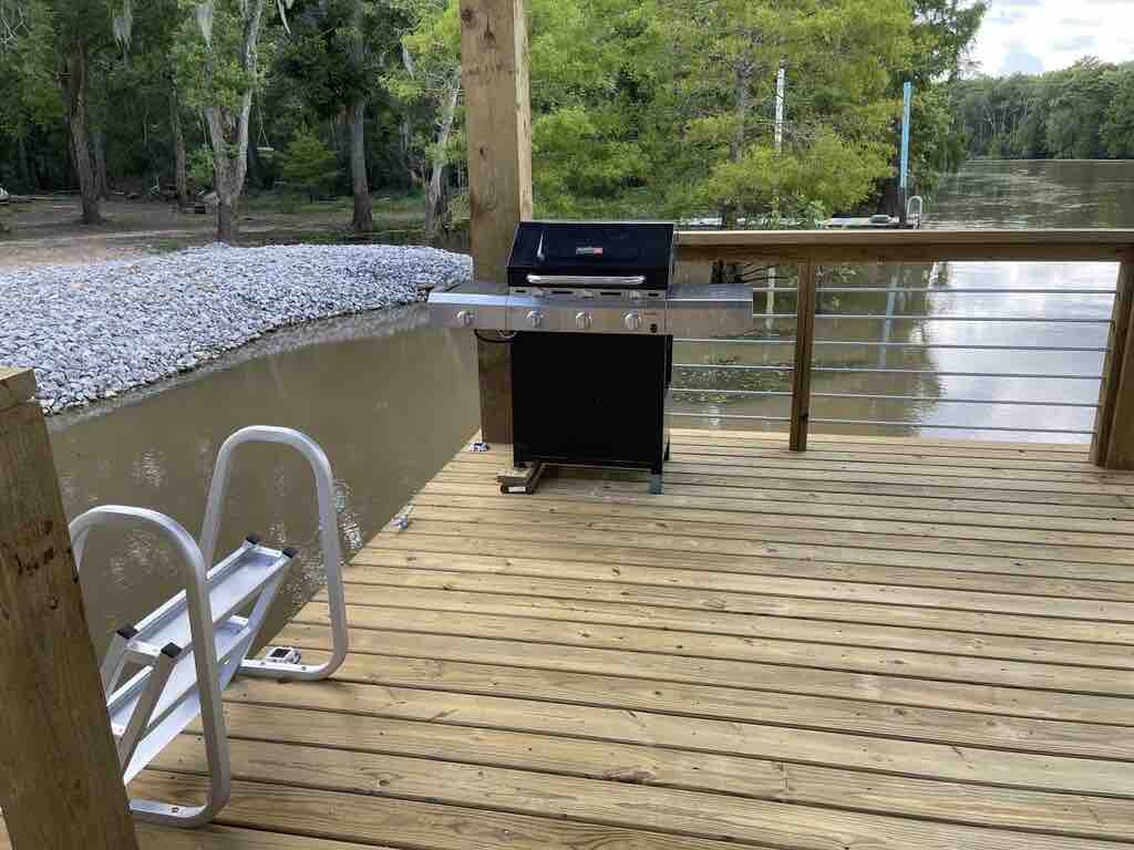 An outdoor deck features a gas grill positioned near the edge, beside a ladder leading to the water. The deck is constructed from wooden planks, surrounded by lush greenery and the flowing river, providing a serene setting for cooking and relaxation.