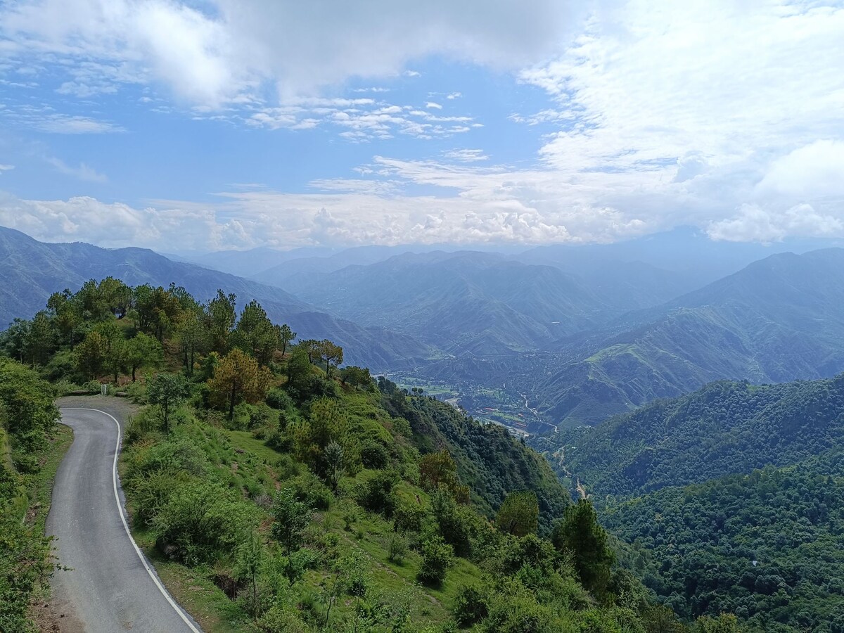 A winding road is nestled between lush green hills, leading toward distant mountain ranges. The landscape is dotted with trees, while valleys stretch beneath a partially cloudy sky, offering expansive views of the natural surroundings.