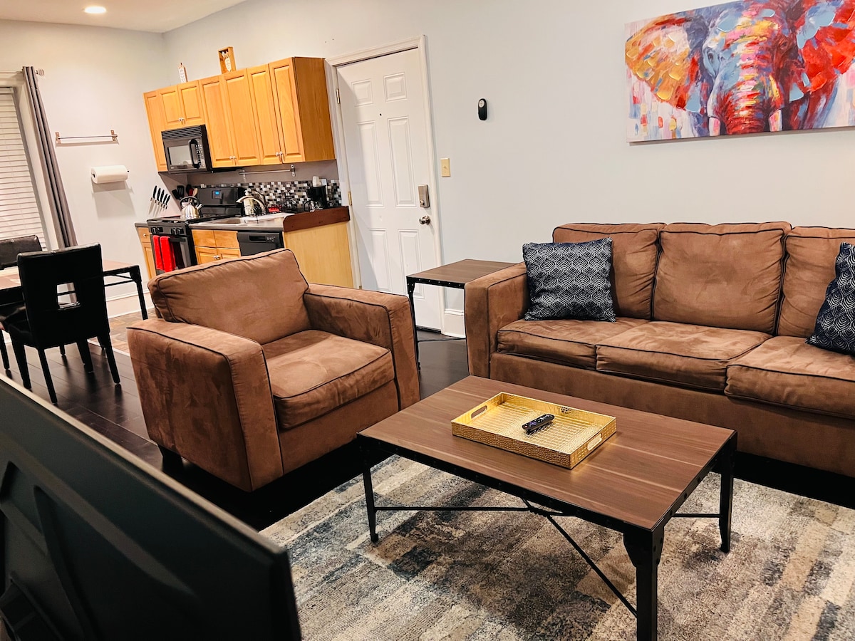 A living area is presented with two brown sofas and a wooden coffee table. An artwork featuring an elephant hangs on the wall, adding color to the neutral interior. A dining area with a black table and chairs is visible in the background.