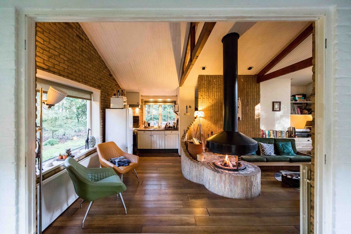 The spacious living area features a central round fireplace and high wooden beams, creating an inviting atmosphere. Large windows allow natural light to fill the room, showcasing comfortable seating and the fully equipped kitchen in the background.