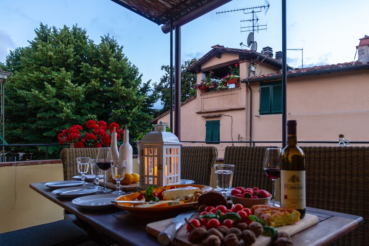 An outdoor terrace features a dining table set with glasses, plates filled with fruits and desserts, and two bottles of wine. Surrounding greenery and neighboring buildings are visible, with red flowers adding color to the scene. Soft lighting is provided by a lantern placed on the table.