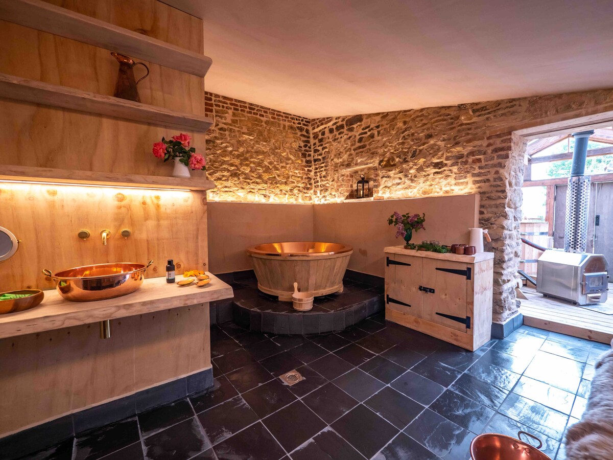 A serene spa room featuring a wooden hot tub set against stone walls. Copper sinks and fixtures complement the wooden vanity, while soft lighting illuminates the space. Outside, views of a private area are available through a glass door, enhancing the tranquil atmosphere.