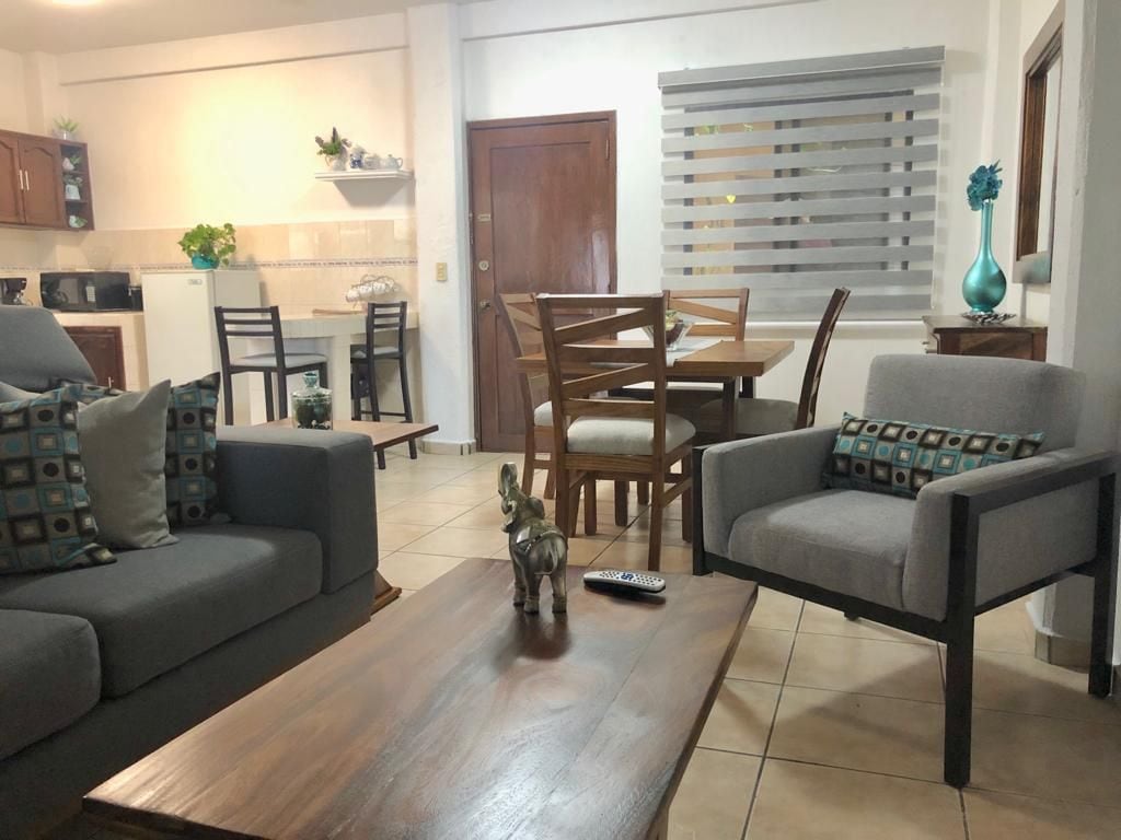 A combined living and dining area showcases a wooden coffee table with decorative accents, surrounded by a sofa and a chair. A dining table for four sits in the background, while a well-equipped kitchen is partially visible. Light filters through a window adorned with horizontal blinds.