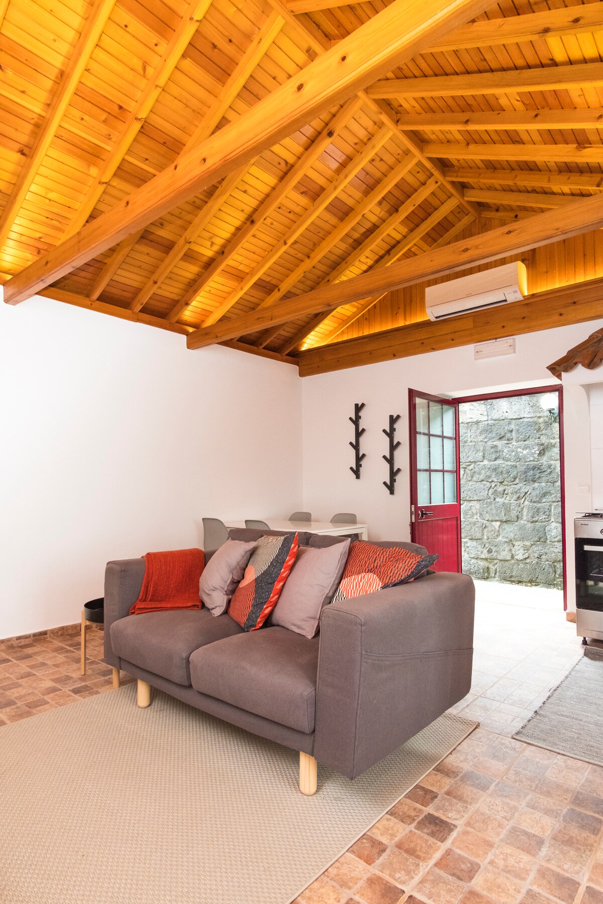 A cozy living area features a gray sofa adorned with colorful cushions, positioned on a textured floor. Exposed wooden beams add warmth to the ceiling. A red door leads to the outside, while a dining area is visible in the background.