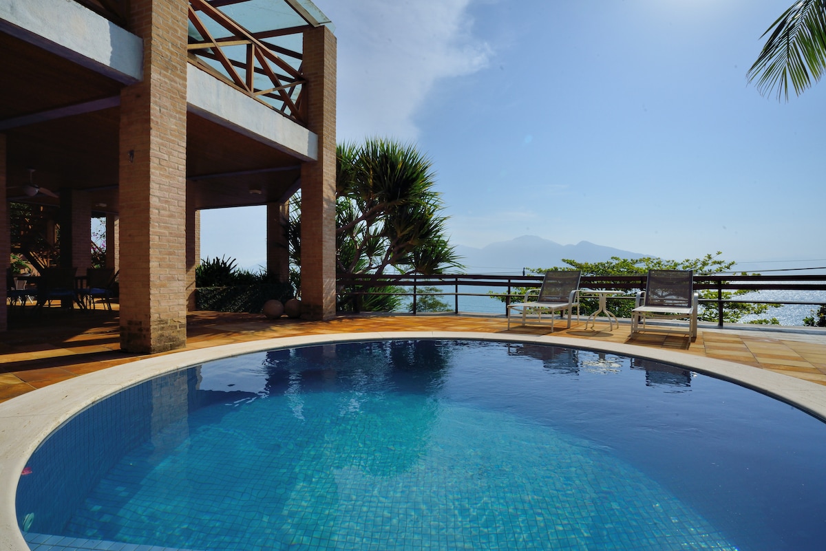 A circular pool glistens under the sun, surrounded by a spacious tiled area featuring two lounge chairs. Lush greenery and distant mountains create a scenic backdrop, while large windows of the adjacent building offer views of the serene surroundings.