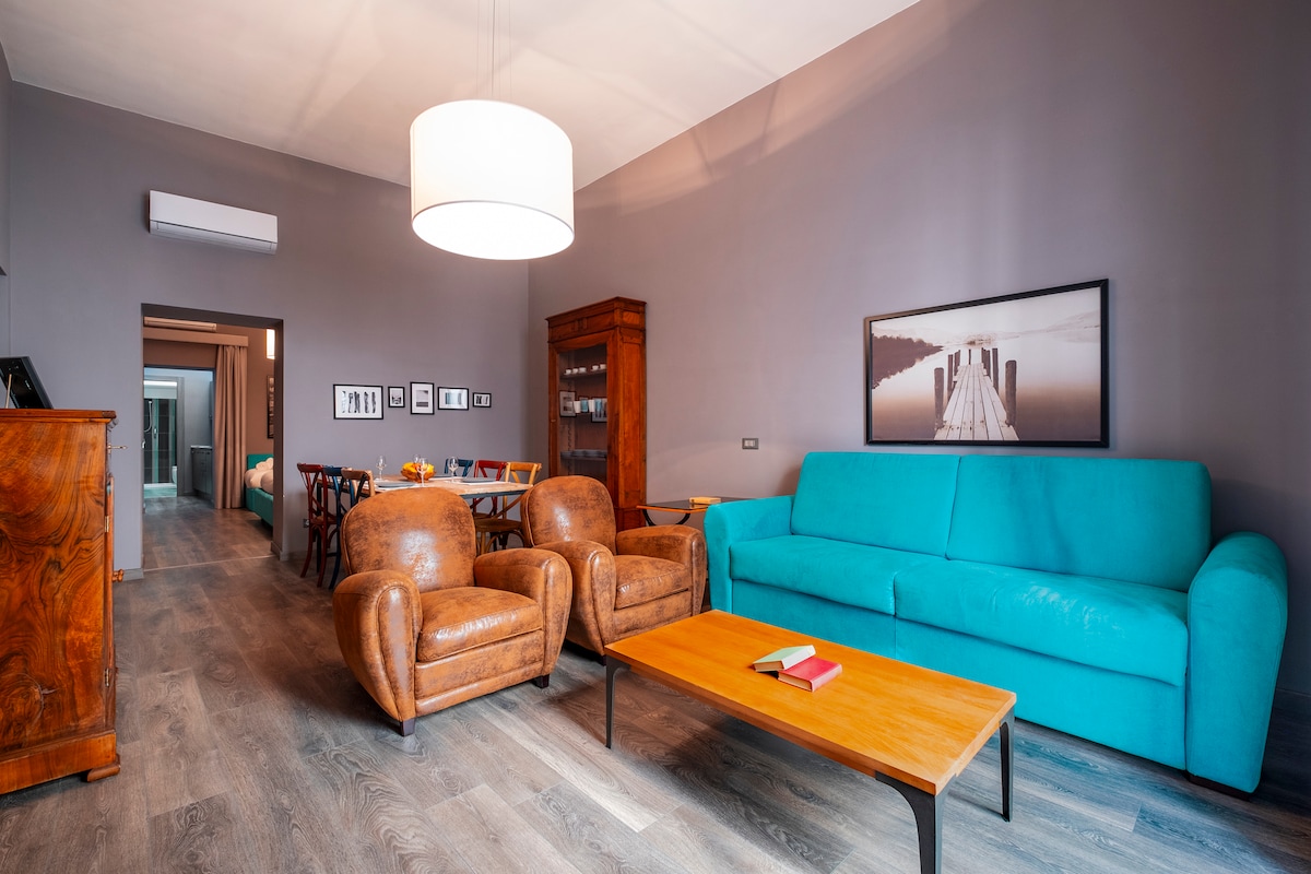 A modern living area features a turquoise sofa and two brown leather armchairs, arranged around a wooden coffee table. A large light fixture hangs above, while artwork depicting a serene landscape adorns the wall. Natural light streams in from the open doorway.