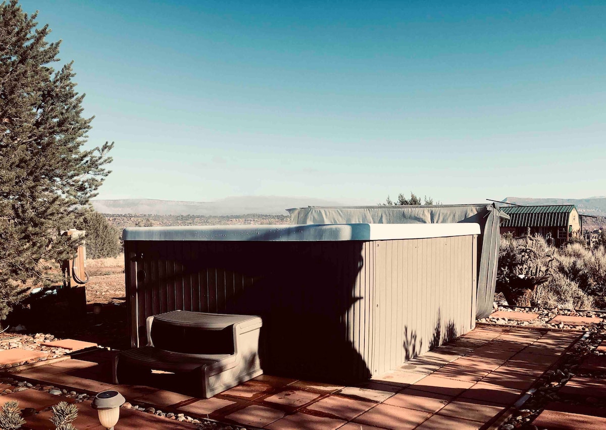 A hot tub is positioned on a stone patio, surrounded by natural terrain. Nearby, a small bench is visible, and distant views of the landscape can be seen against a clear blue sky.