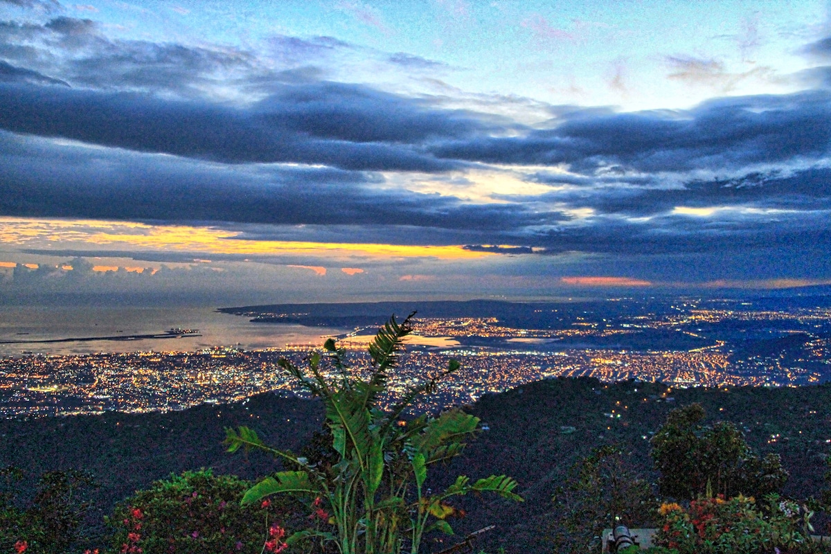 A panoramic view captures the twinkling lights of Kingston below, with the Caribbean Sea visible in the distance. The vibrant sunset paints the sky in soft hues of orange and blue, while lush greenery in the foreground adds a natural element to the serene scene.