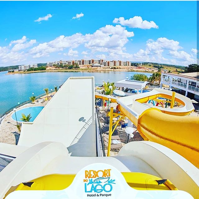 An aerial view captures the vibrant water park at Resort do Lago, highlighting yellow water slides and a serene lake in the background. Lush palm trees dot the landscape, and modern buildings are visible along the shore, providing a sense of relaxation and fun.