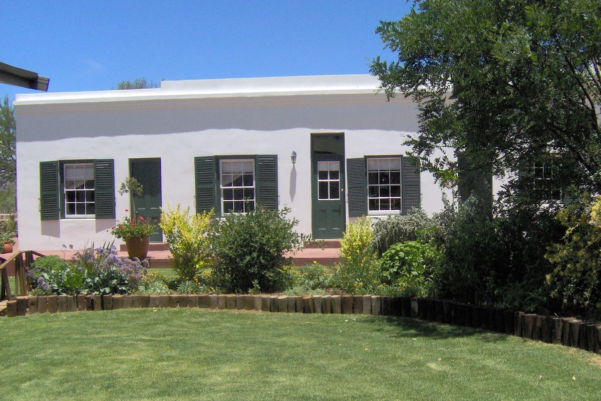 The exterior of a spacious cottage is shown, featuring white walls and green shutters. A covered porch with a door leads into the cottage, surrounded by lush greenery and vibrant flowering plants, creating a welcoming entryway.