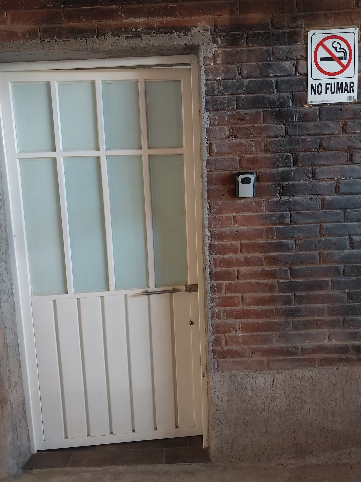 A white, wooden door features multiple glass panes and is set against a textured brick wall. A no smoking sign is displayed above the doorbell, indicating a smoke-free environment. The floor is finished in a smooth, neutral-colored material.