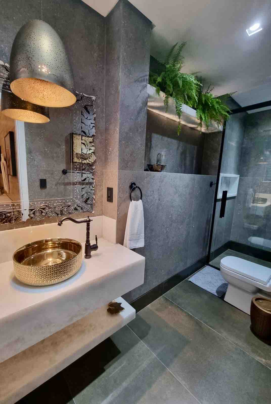The modern bathroom features sleek gray stone walls complemented by a stylish gold basin sink. A frosted glass shower is visible in the corner, while greenery decorates the space. Soft lighting is provided by hanging fixtures, enhancing the overall ambiance.