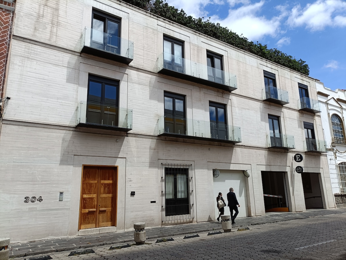 A contemporary building is shown with three levels and several large glass balconies. The facade features a light-colored finish, while the entrance is framed by a wooden door. The street in front is lined with cobblestones, and people are seen walking by.