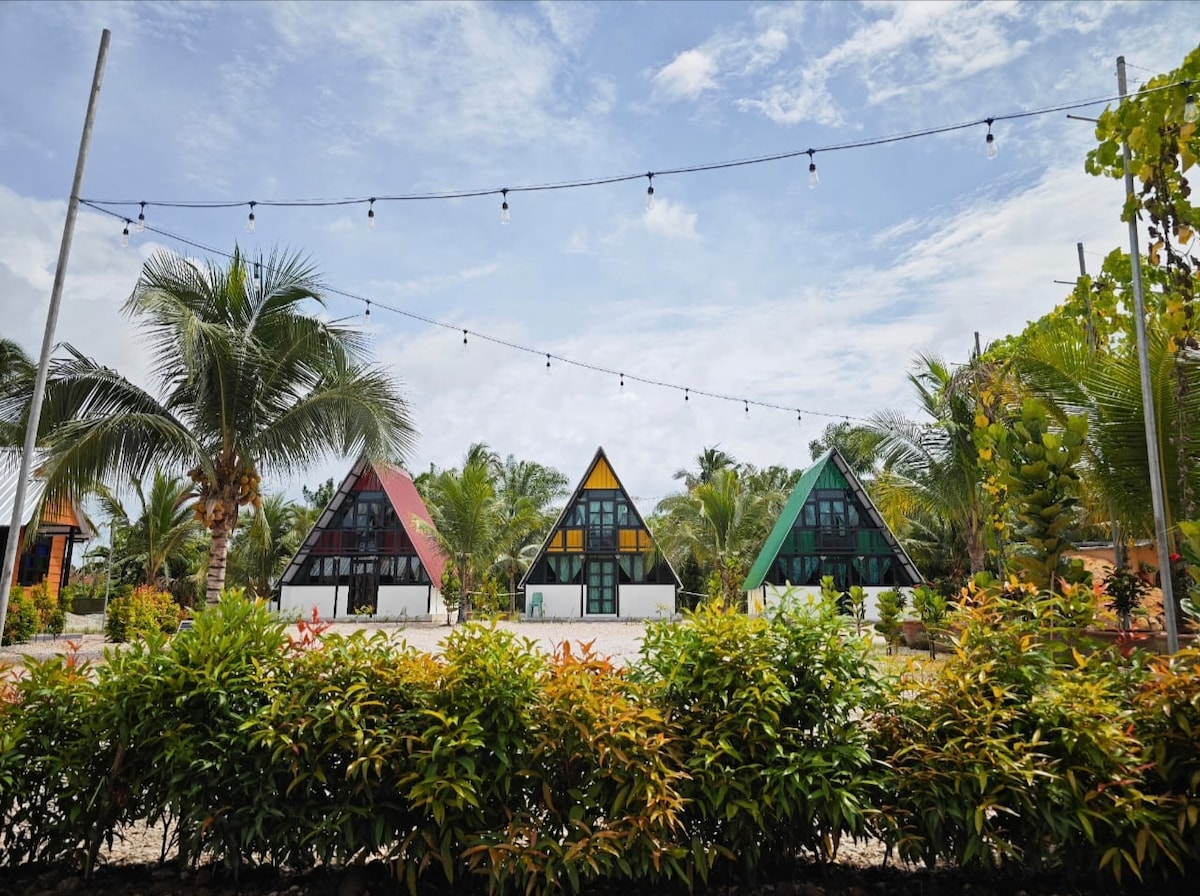 Three A-frame villas are visible, each featuring distinct colored roofs in red, yellow, and green. Surrounding greenery includes palm trees and flowering plants. String lights are suspended above the area, enhancing the outdoor space. A well-maintained garden frames the villas, creating a serene atmosphere.