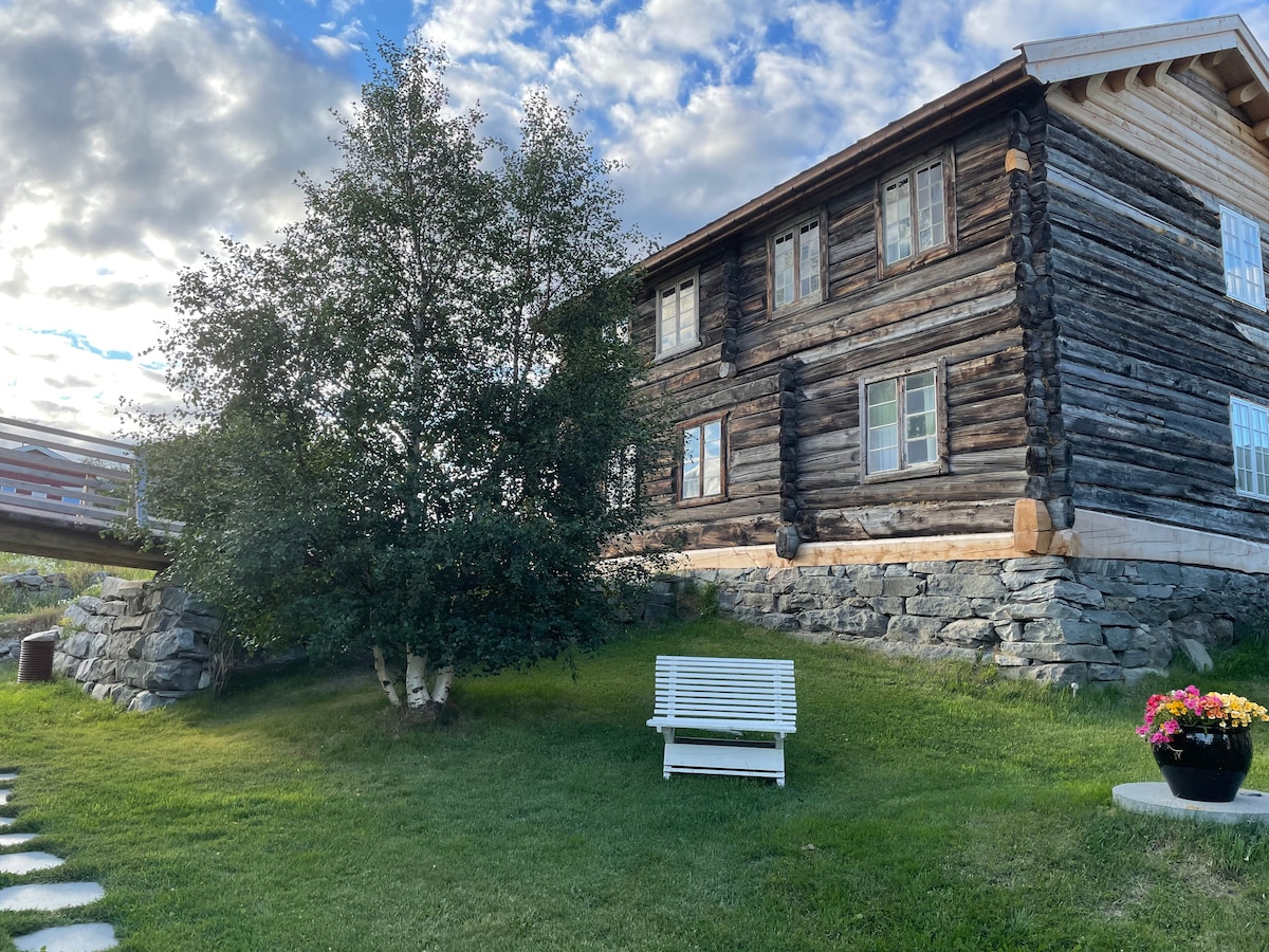 A historic wooden building, characterized by its traditional logs, stands on a gently sloping green landscape. A white bench is positioned on the lawn, surrounded by flowering plants in a pot. The sky is partly cloudy, adding a serene atmosphere.