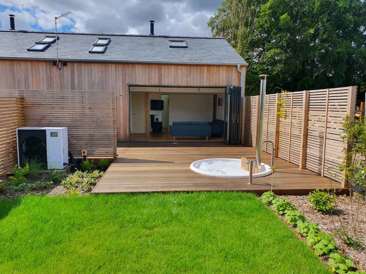 A wooden deck surrounded by green grass features a wood-burning hot tub. The exterior of the converted barn is seen in the background, with large sliding doors leading into a living area. Shrubs and plants provide a natural border to the space.