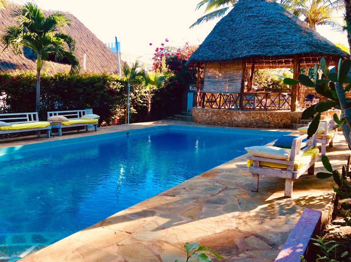 A tranquil outdoor area features a clear swimming pool surrounded by a stone deck. Sun loungers are positioned at the pool's edge, and a thatched-roof gazebo is visible in the background, framed by lush greenery and vibrant flowers.
