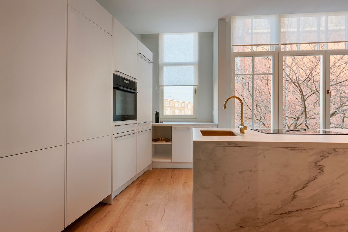 A modern kitchen showcases white cabinetry, equipped with sleek appliances and a marble island that enhances the minimalist design. Large windows allow for natural light to fill the space, offering a view of the outside cherry tree landscape.