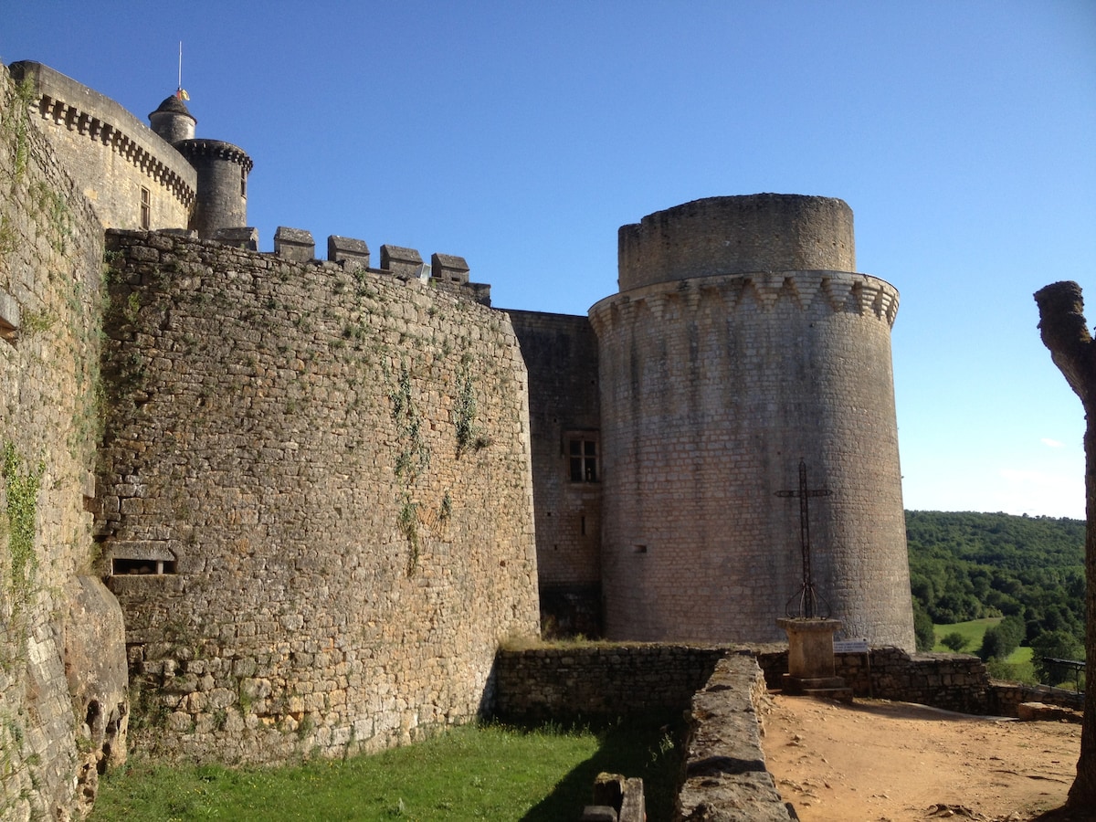 A historic stone fortress stands prominently with thick walls and a round tower. Lush green hills extend in the background under a clear blue sky, providing a serene backdrop. The surrounding area features a sandy path leading up to the structure, creating an inviting atmosphere.